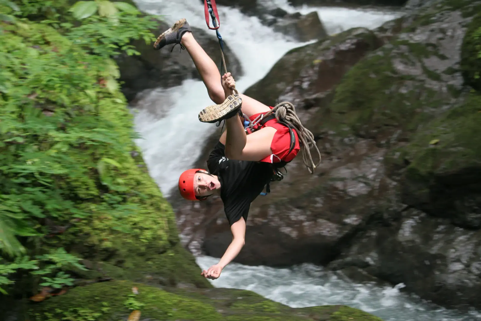 Persona con casco rojo colgando de cabeza en rappel junto a una catarata en la selva