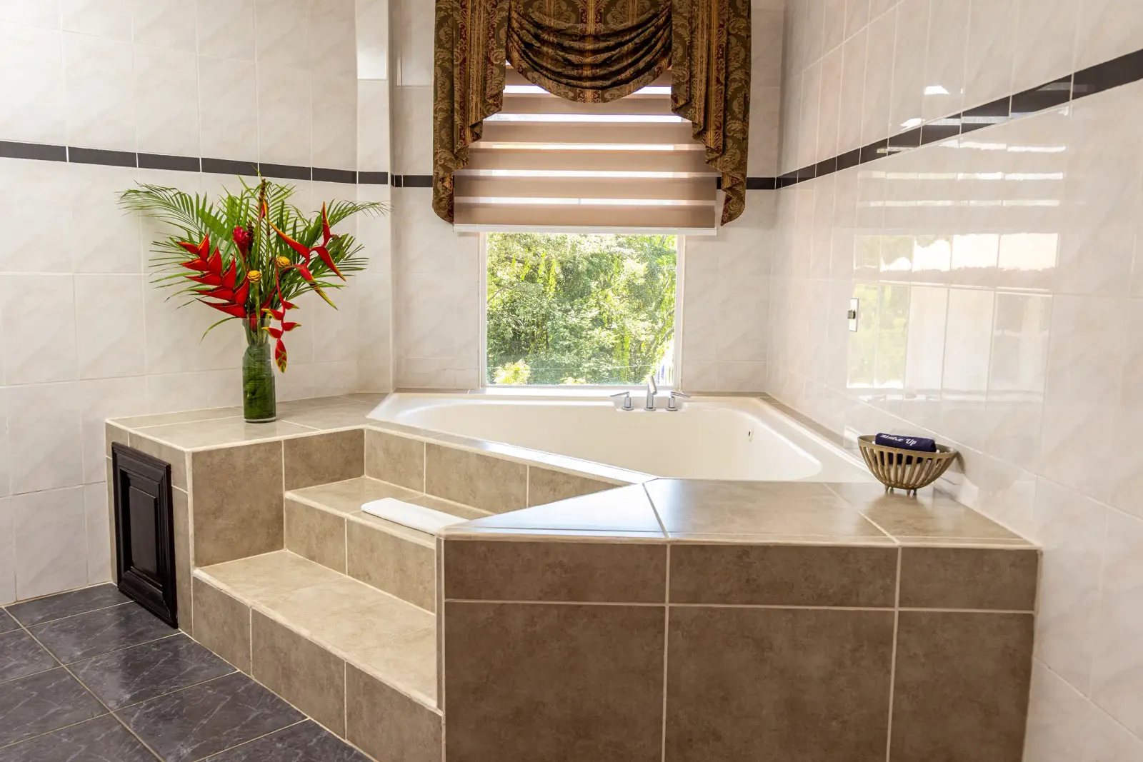 Family Suite bathroom with a tiled jacuzzi tub under a window and a vase of heliconias