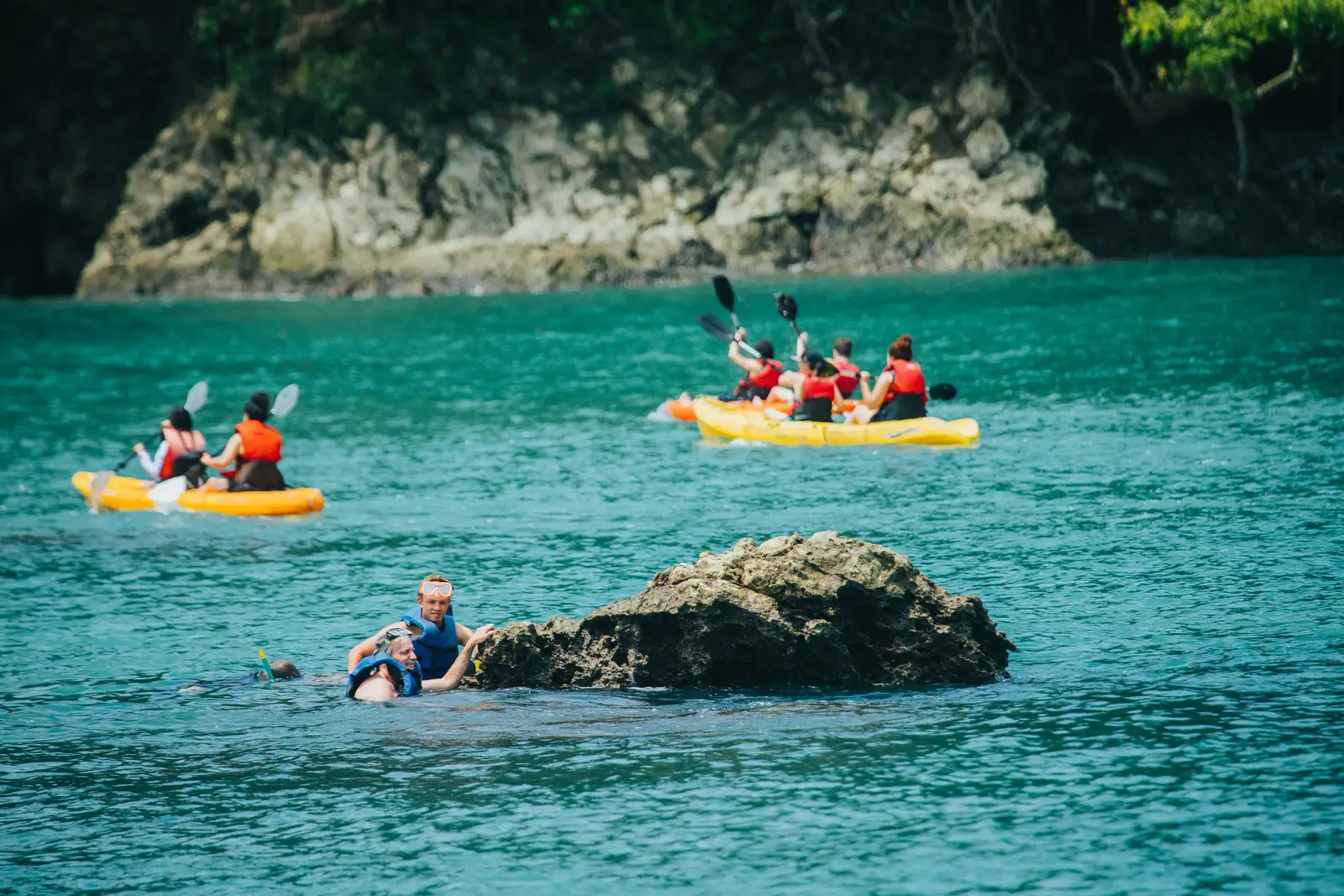 Personas en kayaks amarillos remando cerca de rocas bajo un acantilado con bosque