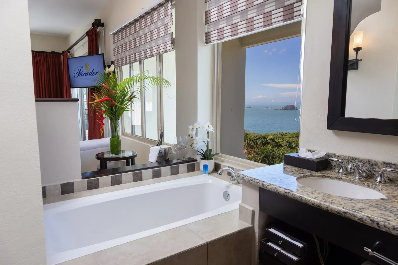 Soaking tub under a window with an ocean view, beside a granite vanity with an orchid