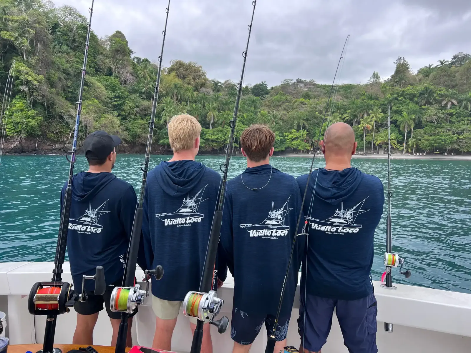 Four anglers in Vuelto Loco hoodies at the stern with rods, facing a forested shore