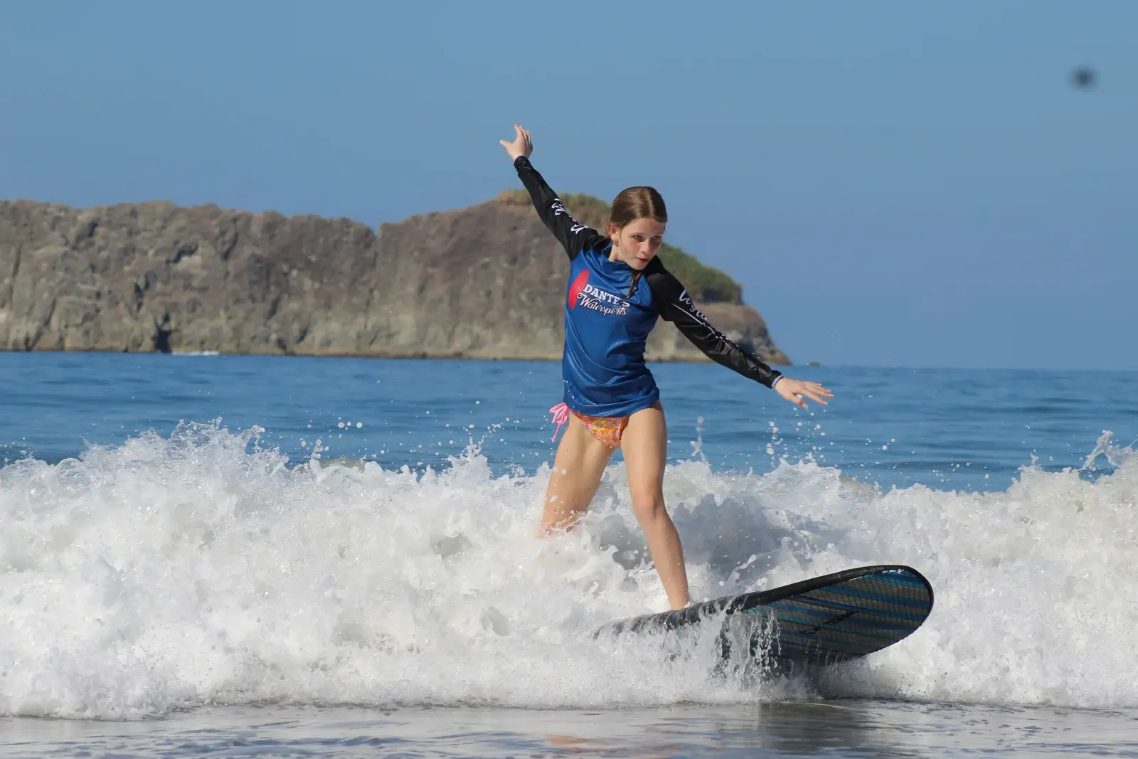 Niña con licra de surf montando una ola pequeña, con un promontorio rocoso al fondo