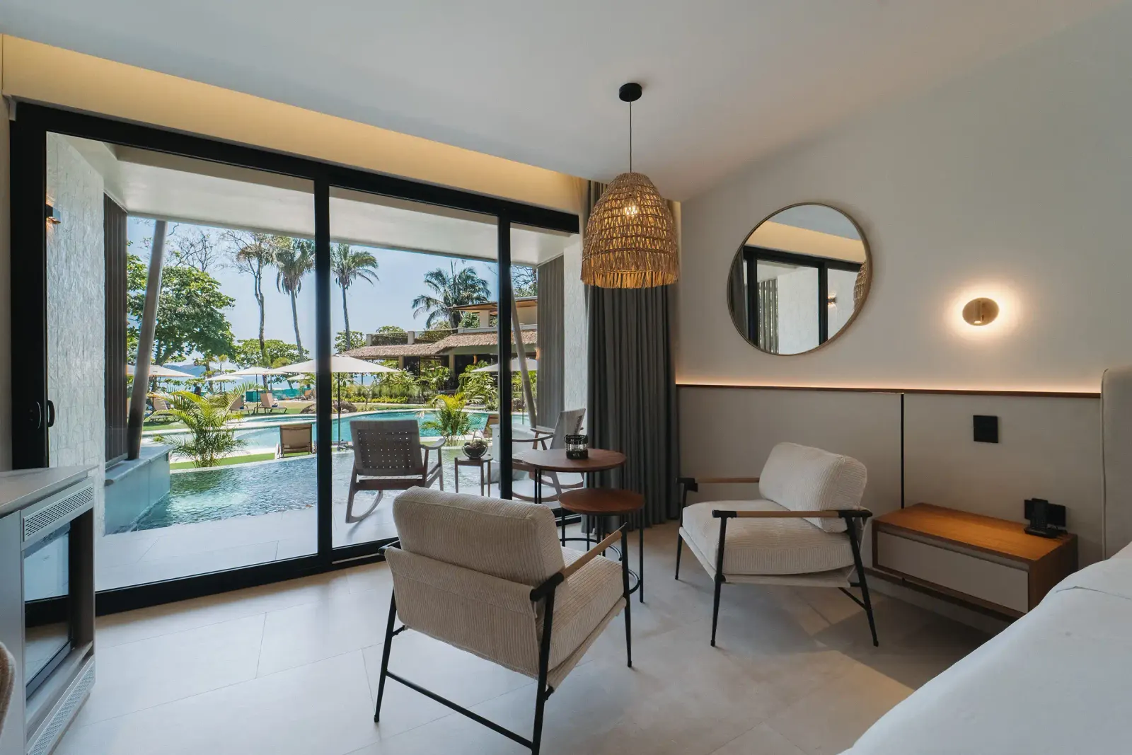 Habitación con puertas corredizas de vidrio abiertas a una terraza junto a la piscina y palmeras afuera, una lámpara colgante tejida, un espejo redondo y una sala con sillas beige y mesitas.