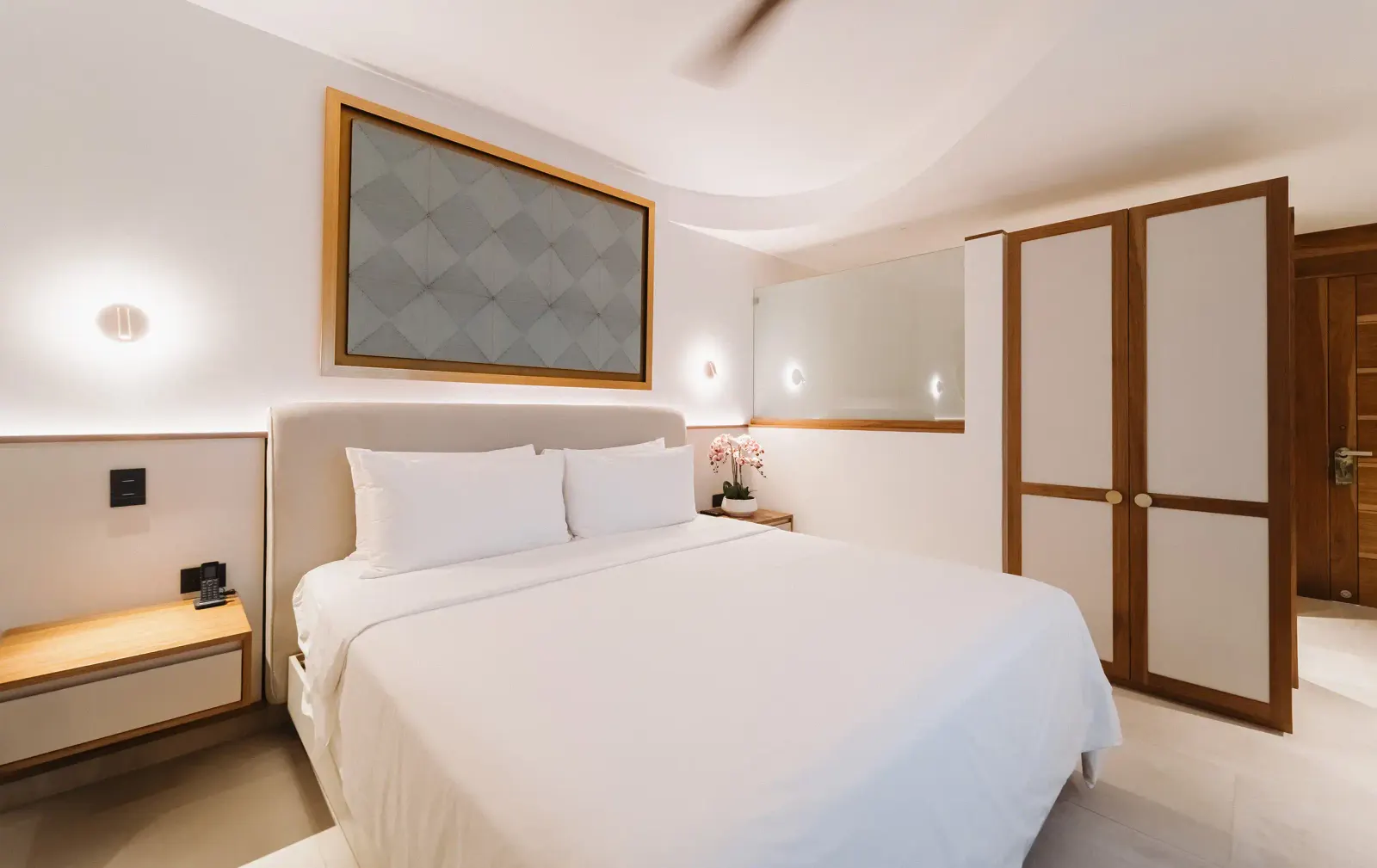 Habitación con cama king de sábanas blancas y dos mesas de noche de madera contra una pared de cabecera tapizada gris.