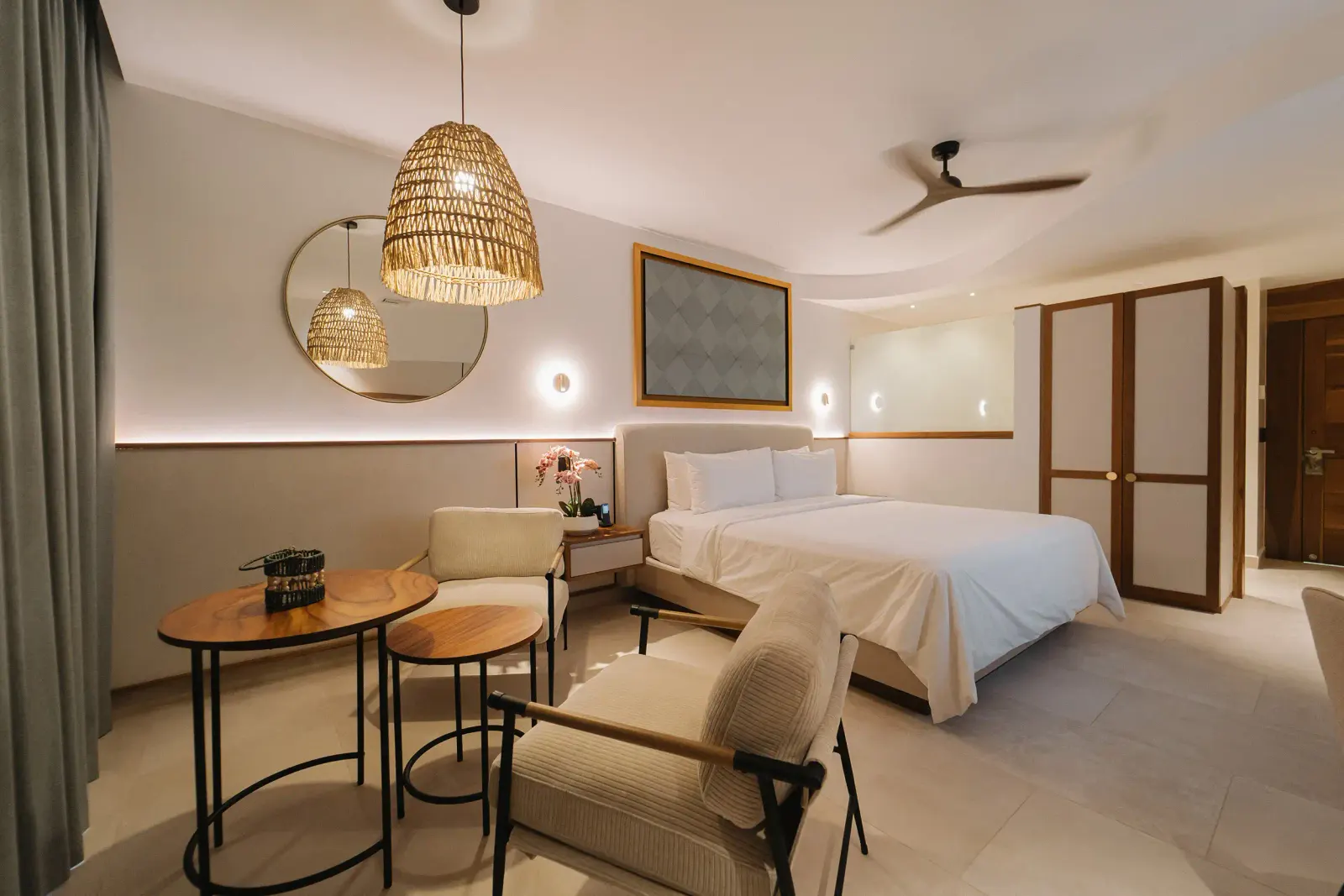 Habitación con dos camas blancas, lámparas colgantes tejidas, un espejo redondo y un ropero de madera a la derecha.
