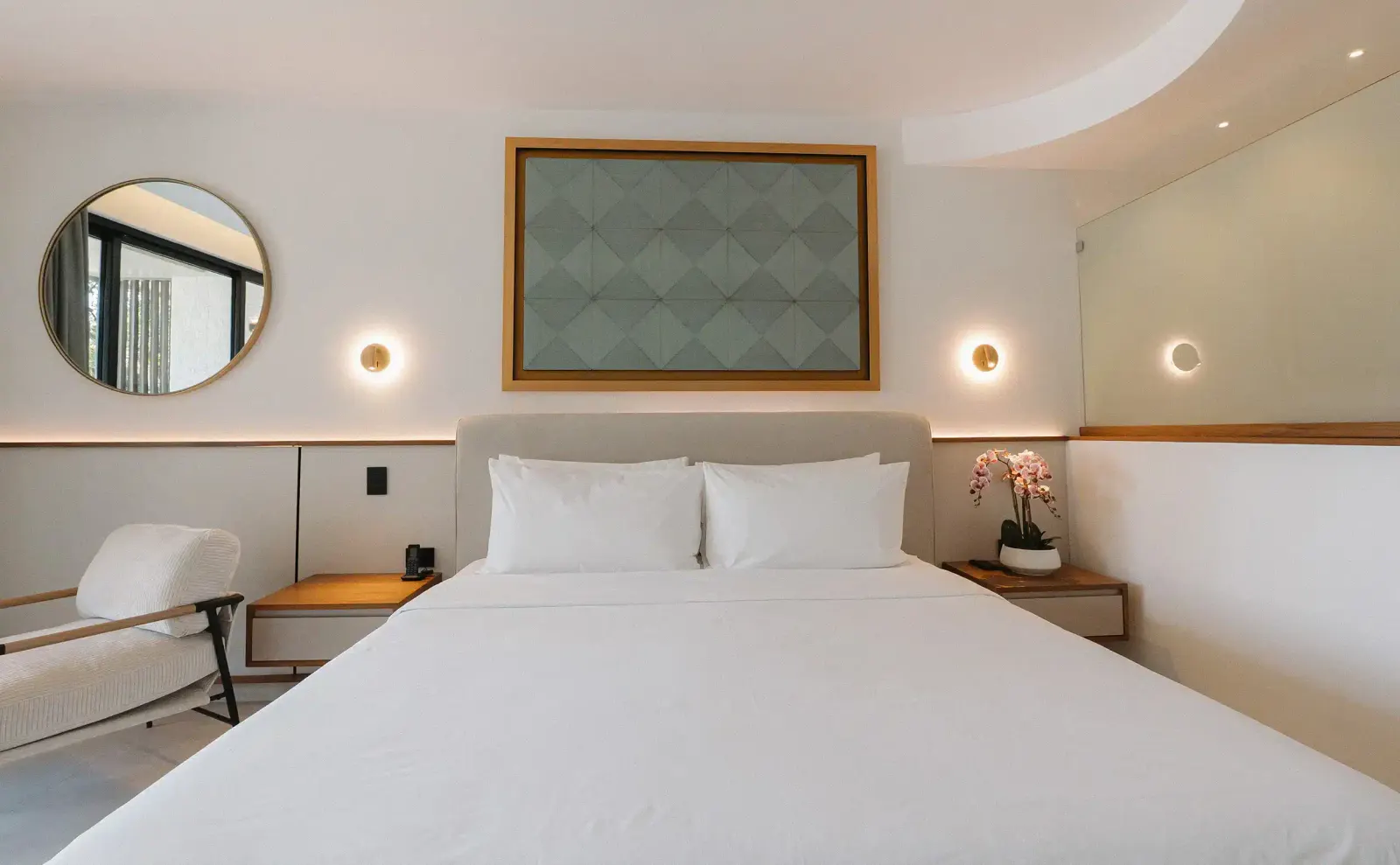 Vista amplia de un dormitorio luminoso con una cama blanca grande, cabecera tapizada, un espejo redondo en la pared izquierda y arte geométrico sobre la cama.