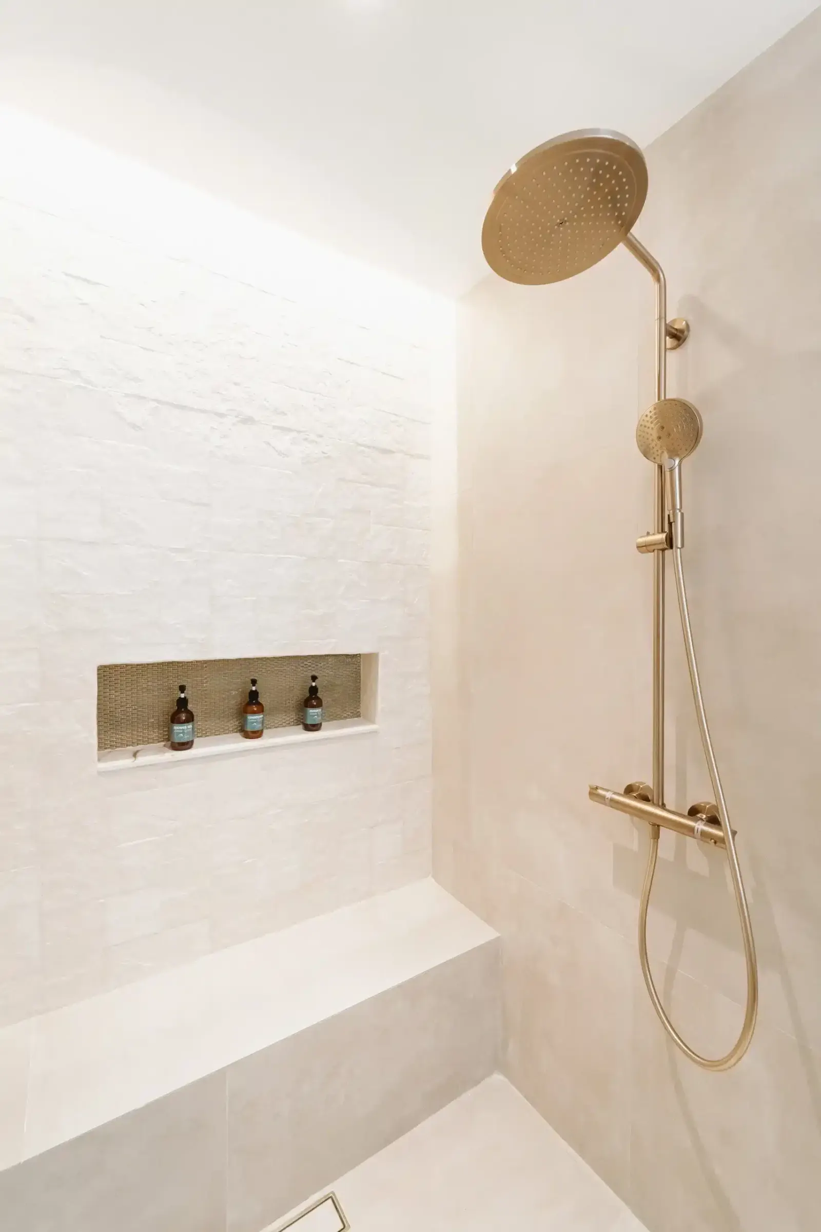 Ducha con acabados de bronce, una regadera tipo lluvia grande y una de mano, paredes de azulejo blanco y gris y una repisa empotrada.