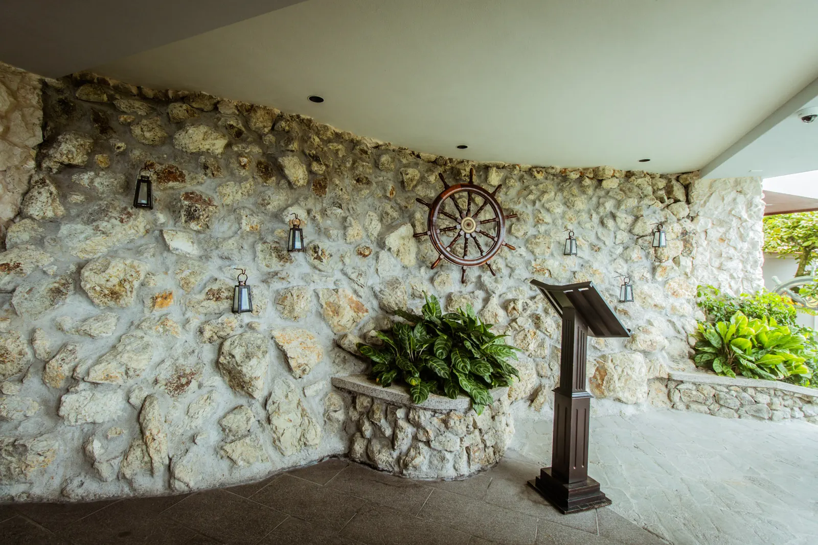 A curved stone wall with a ship's wheel and lanterns at the entrance to La Fragata, a host stand and potted greenery