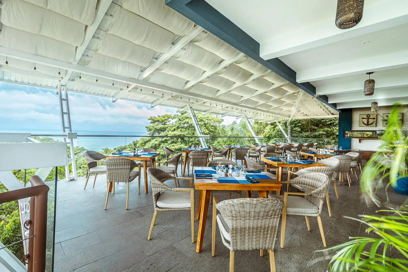 La Fragata's terrace from the far corner, set tables under the fabric shades, a palm frond in front and the ocean through the trees