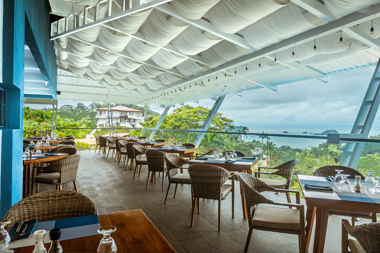 La Fragata's open-air terrace under white fabric shades, wicker chairs at set tables, the Pacific and its rocky islands beyond a glass railing