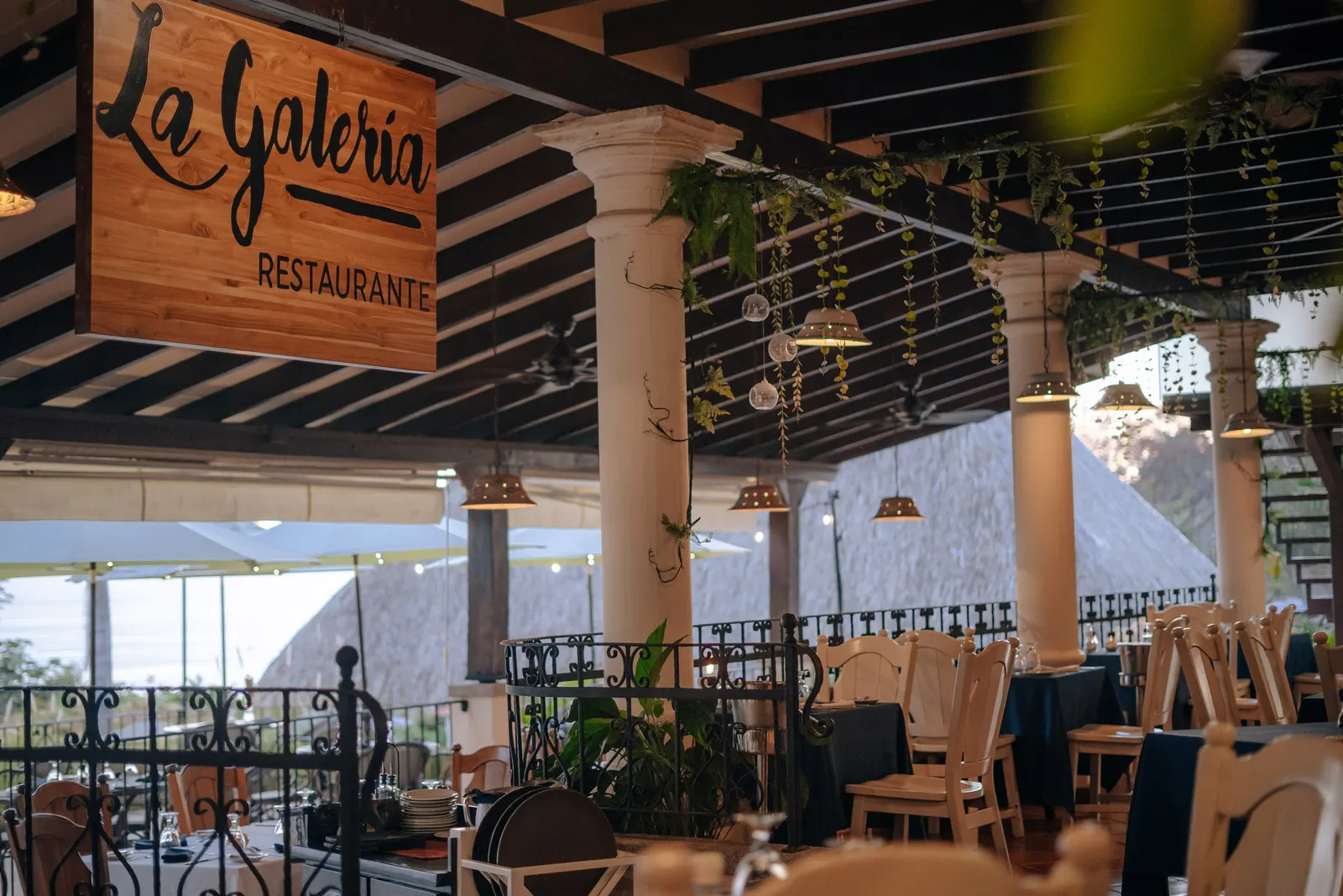 La Galería restaurant terrace at dusk, set tables under hanging plants, ocean beyond