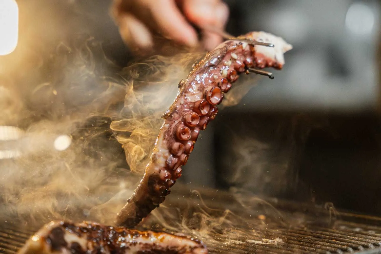 An octopus tentacle lifted from the grill with tongs, smoke rising