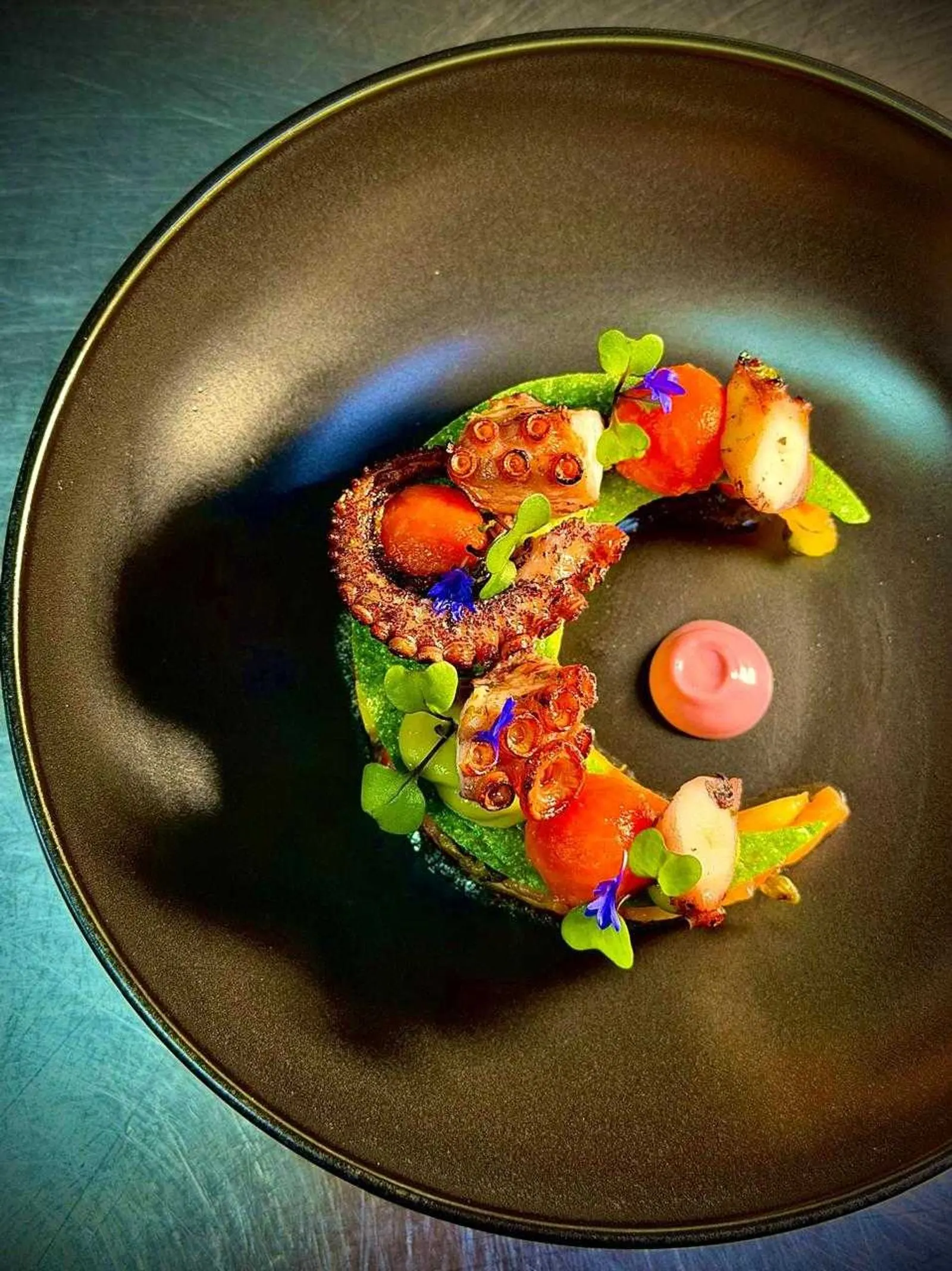 Grilled octopus arranged in a crescent with tomatoes, sprouts and edible flowers on a dark plate