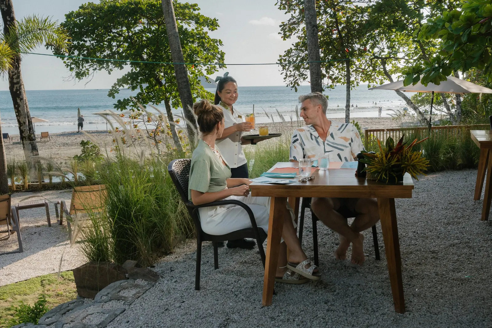 Una mesera lleva bebidas a una pareja en una mesa al aire libre de Yemaya, con la playa y el Pacífico detrás