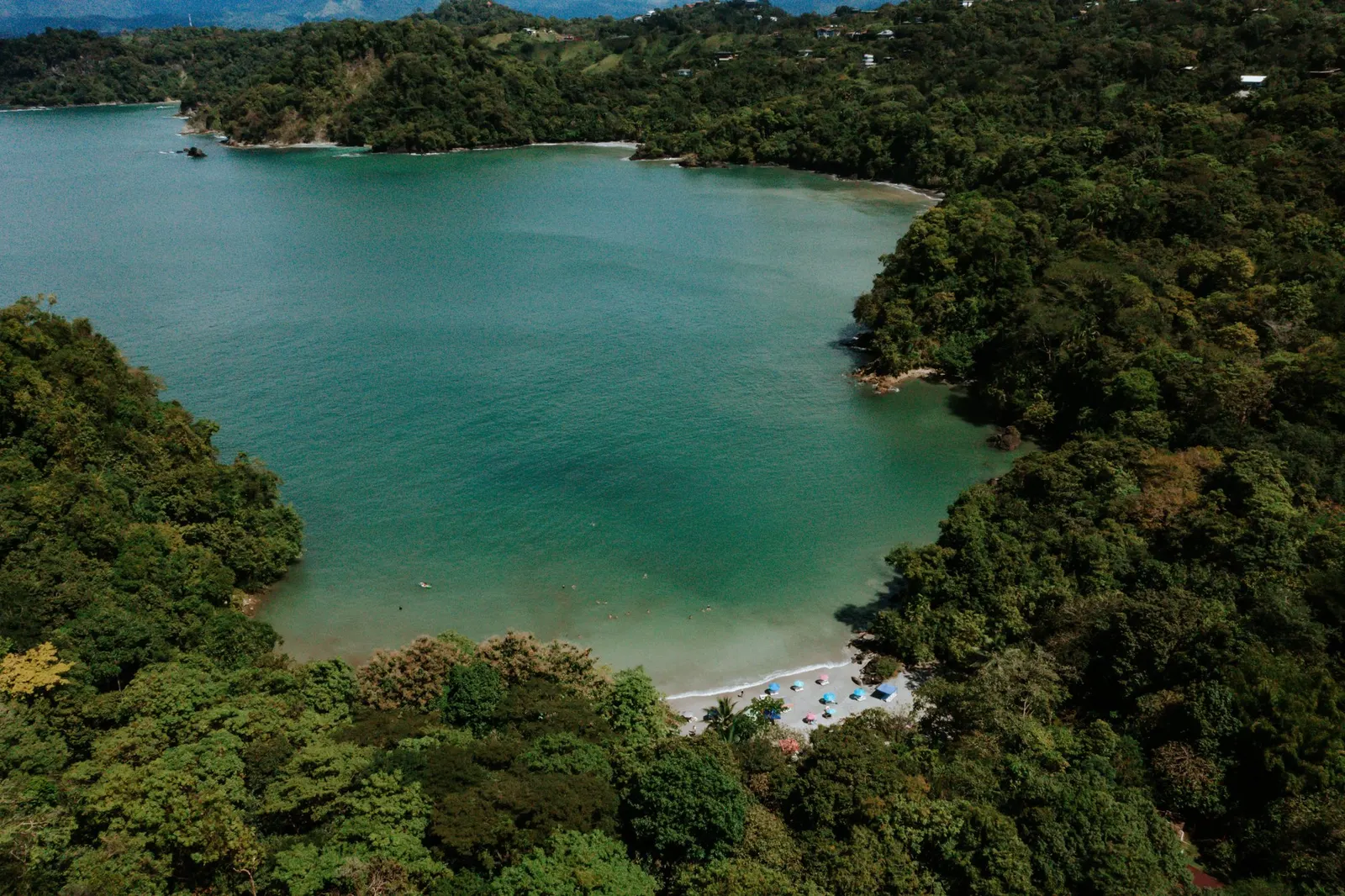 Vista aérea con dron de una caleta boscosa en Manuel Antonio, con agua turquesa, playa de arena blanca y sombrillas de colores
