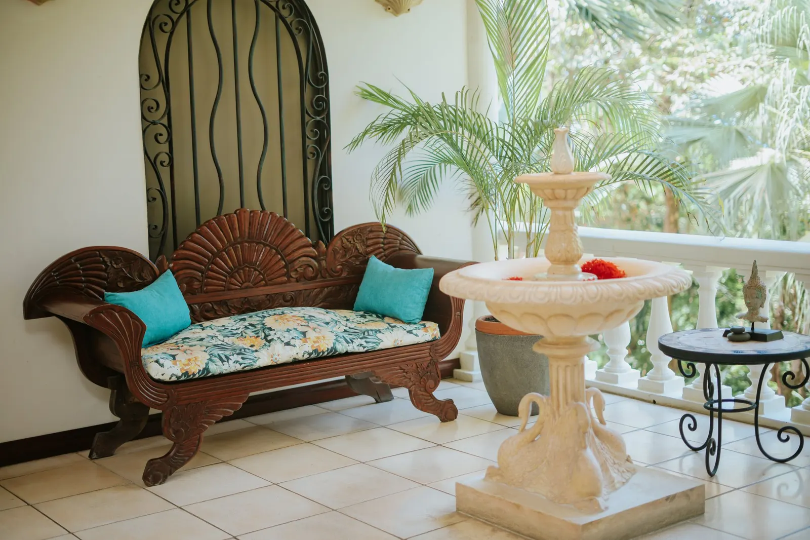 Vista amplia de una banca de madera tallada con almohadas turquesa y un cojín floral junto a una fuente de piedra con pedestal de cisne y flores rojas de ixora flotando, enmarcada por una palmera en maceta y la baranda de un balcón con vista a la selva