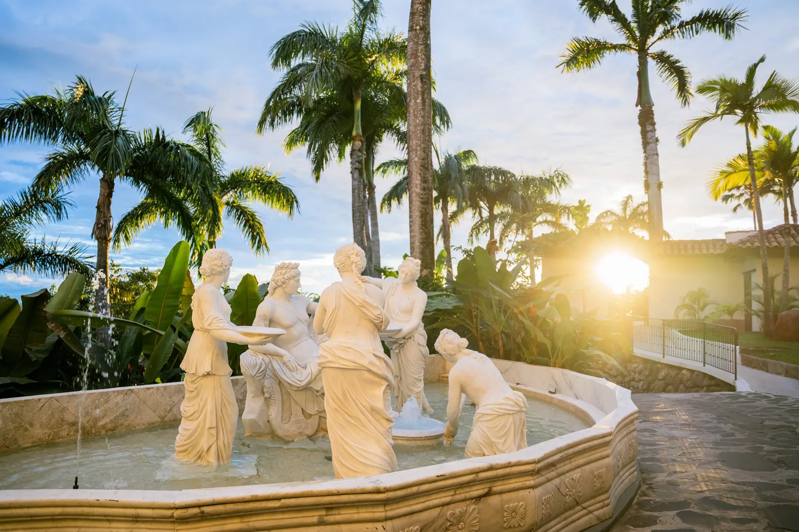 Grupo de estatuas de mármol en una fuente al atardecer, una figura inclinada hacia el agua en el borde de la fuente y el sol brillando a través de un portón del jardín al fondo