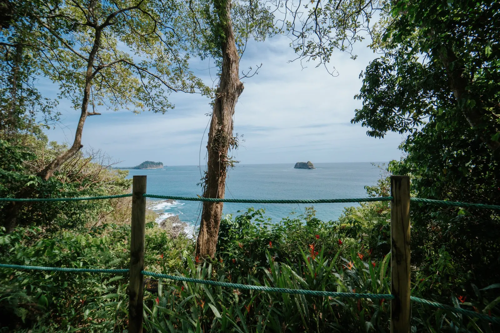 Vista cercana entre troncos y una cerca de mecate en un mirador del jardín, con dos islotes rocosos en el océano y flores de heliconia roja abajo