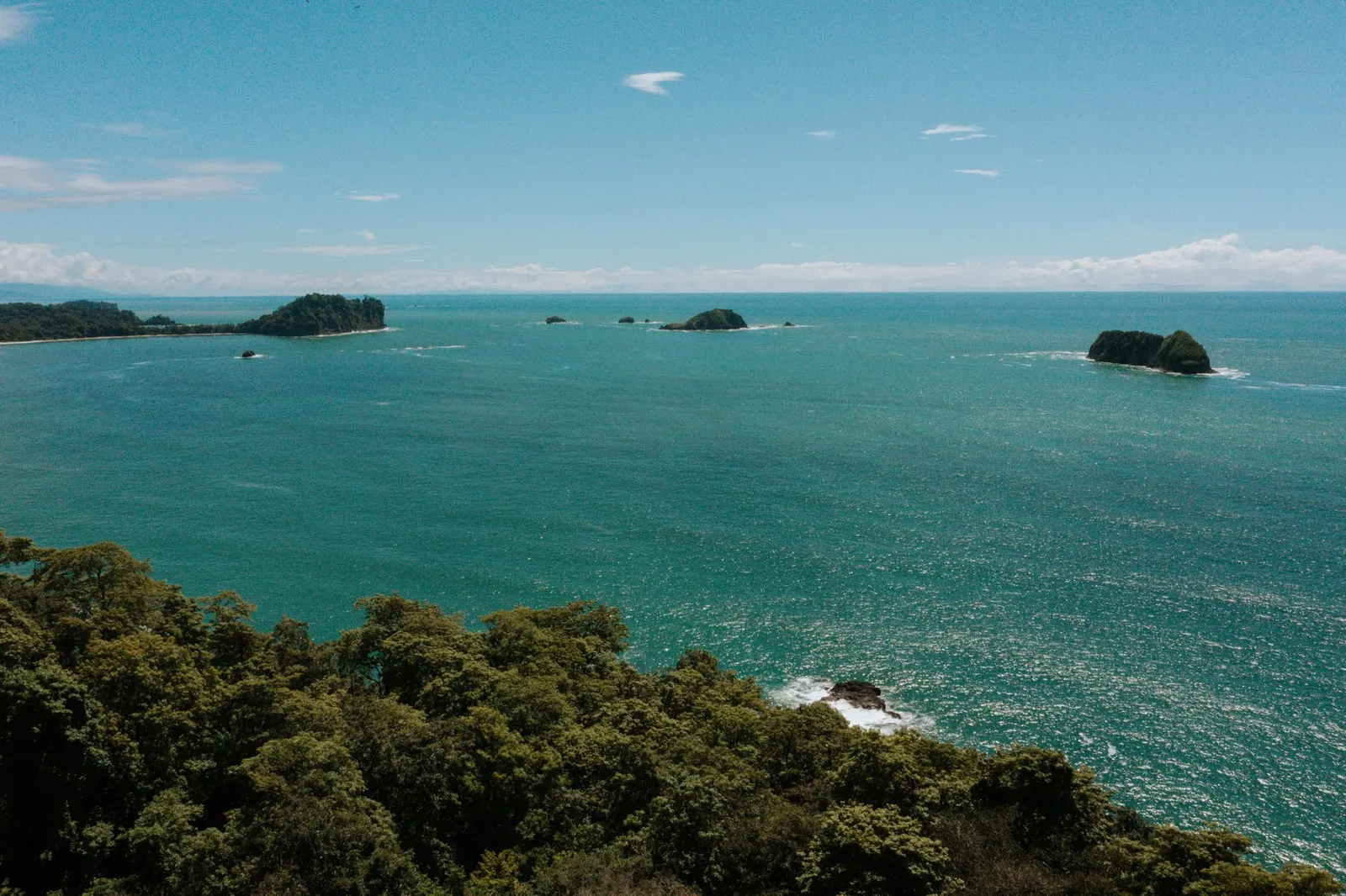 Vista elevada de día sobre las copas del bosque tropical hacia el océano Pacífico turquesa, con varios islotes rocosos frente a la costa