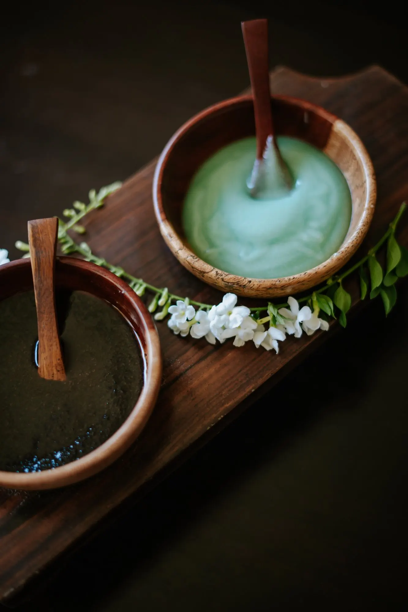 Dos cuencos de madera con mascarillas de arcilla verde y oscura con espátulas, sobre madera oscura con una guirnalda de flores de jazmín