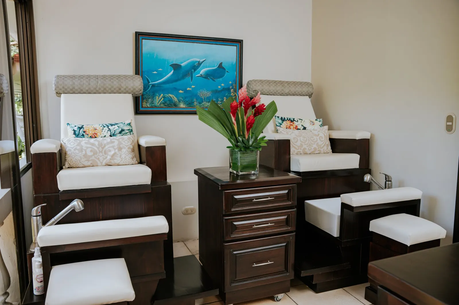 Primer plano de los dos sillones de pedicura color crema con almohadas florales, centrado en un florero con flores de jengibre rojo sobre el mueble de madera oscura que está entre ellos