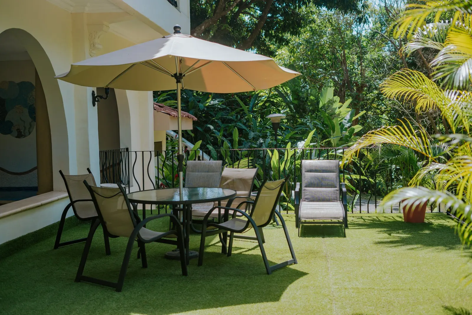 Una terraza al aire libre con una mesa redonda, sillas de lona, una silla reclinable y una sombrilla beige abierta, con el borde de una piscina de mosaico junto a un arco blanco y la selva más allá