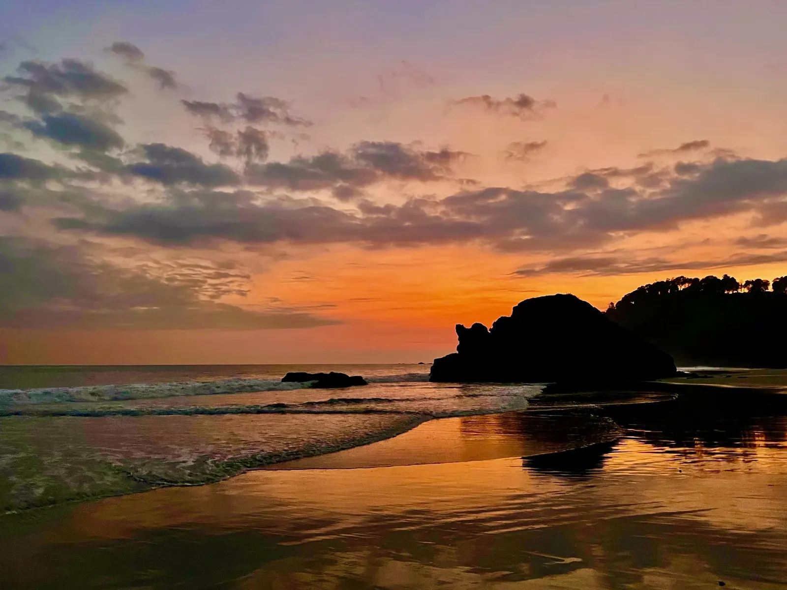 Un amplio atardecer naranja y morado sobre el océano, con una formación rocosa oscura recortada contra la arena mojada que refleja el cielo