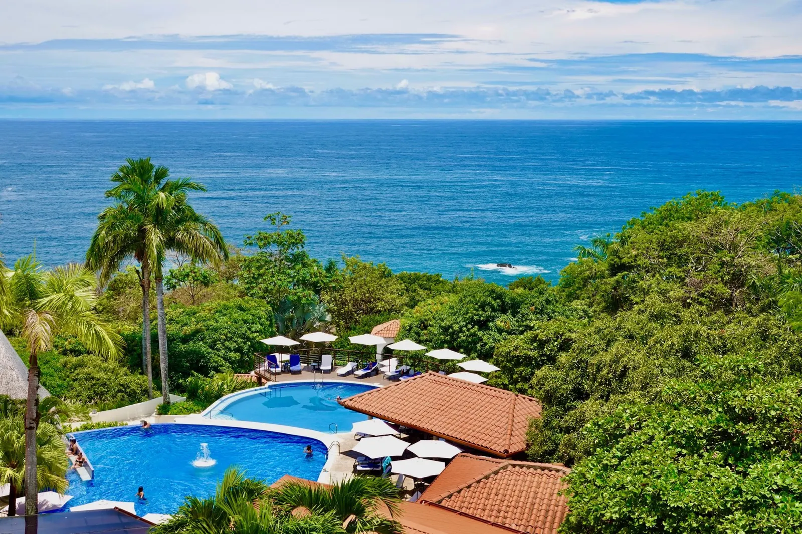 Vista aérea amplia de la piscina del resort y el área de tumbonas que la rodea, con el océano extendiéndose hasta el horizonte