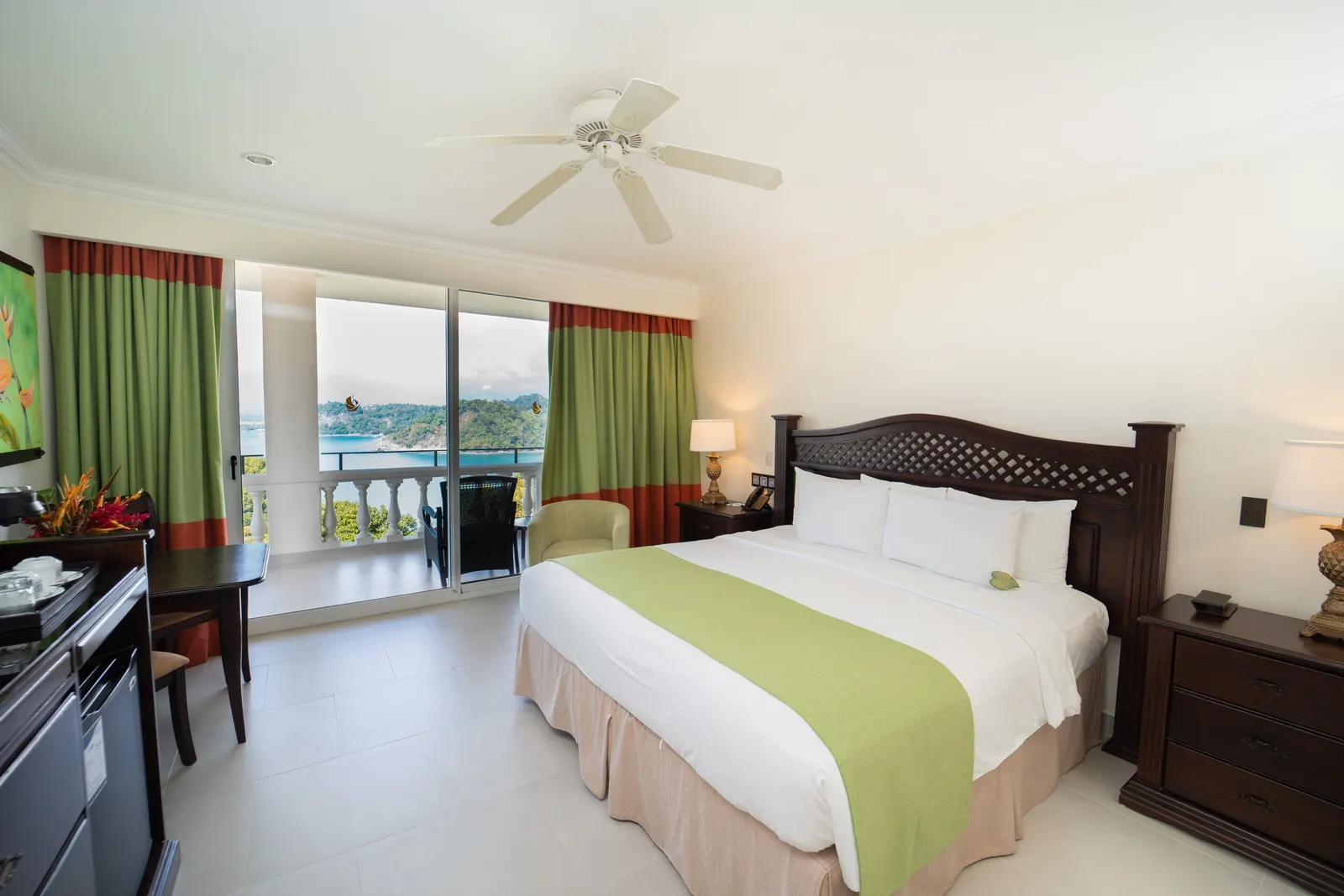 King bed with a green runner and a leaf-shaped welcome card on the pillow, curtains drawn back from a sliding door to a balcony over the ocean and the forested coast