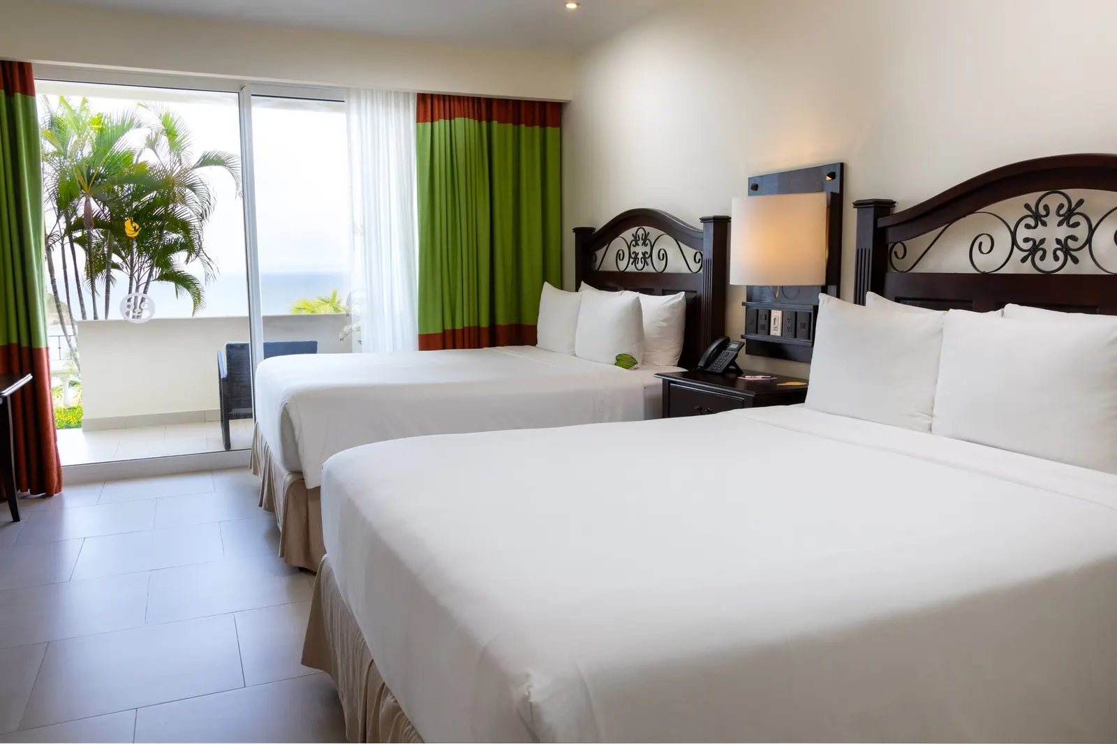 Two beds with carved dark wood headboards beside a sliding glass door with a glimpse of palms and the ocean