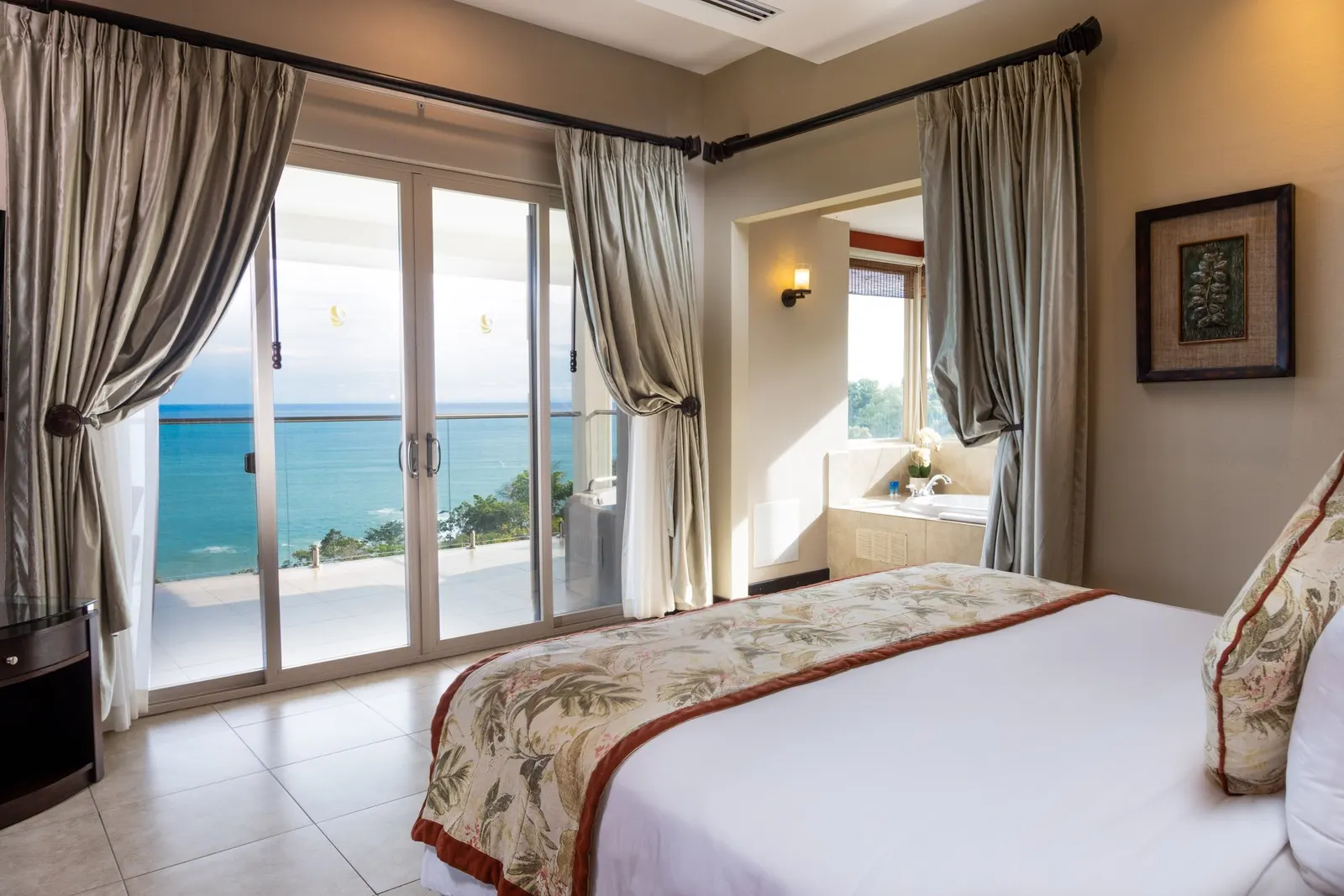 King bed with a floral runner facing open sliding glass doors to a terrace and the ocean, a soaking tub in the window alcove beyond