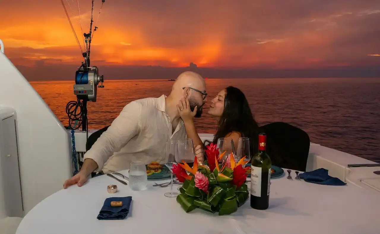 A couple share a kiss at a dinner table set on the deck at sunset, with wine and a tropical flower centerpiece