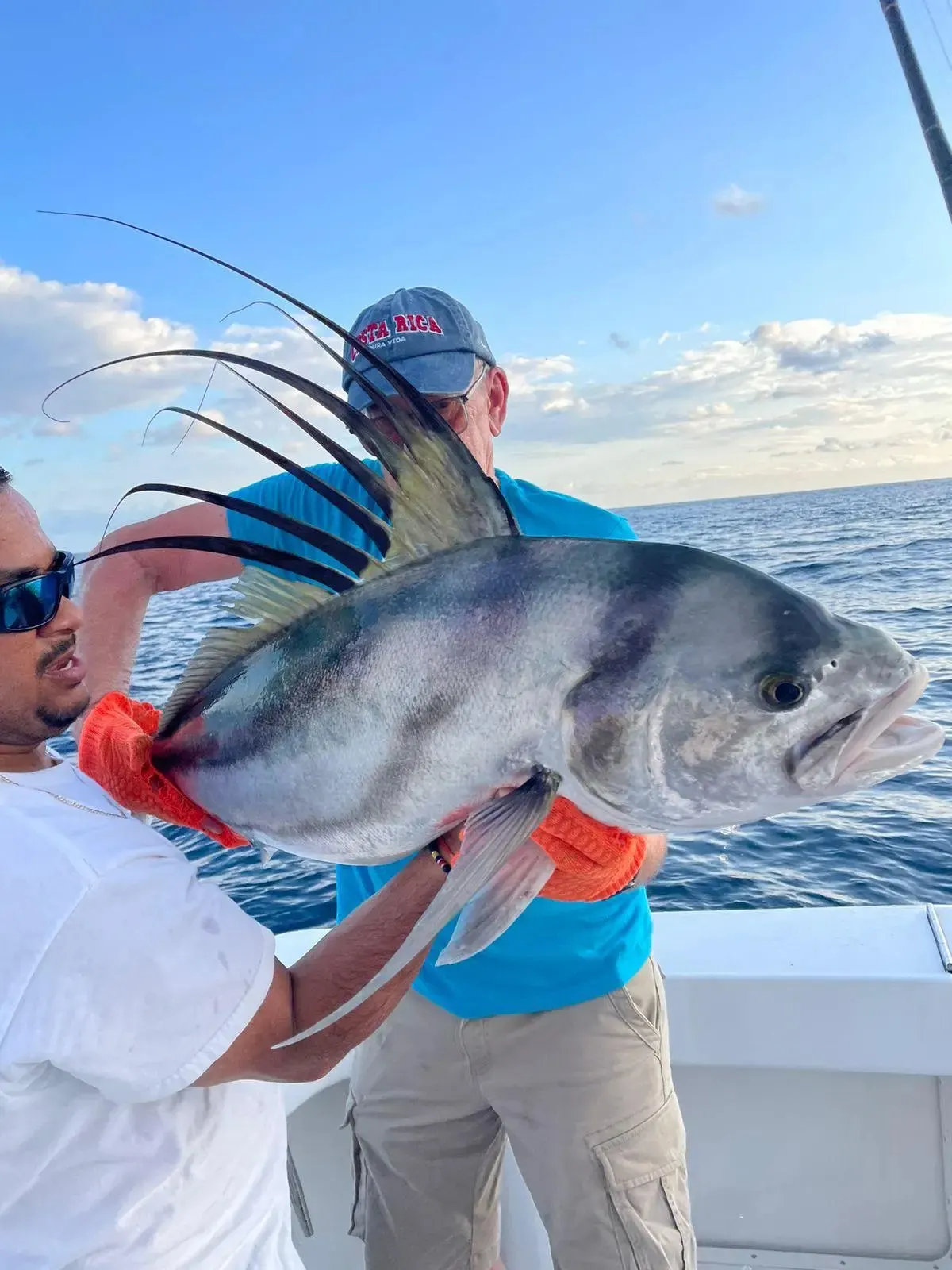 Two anglers on the boat hold up a roosterfish, its long comb of dorsal spines raised against a blue sky