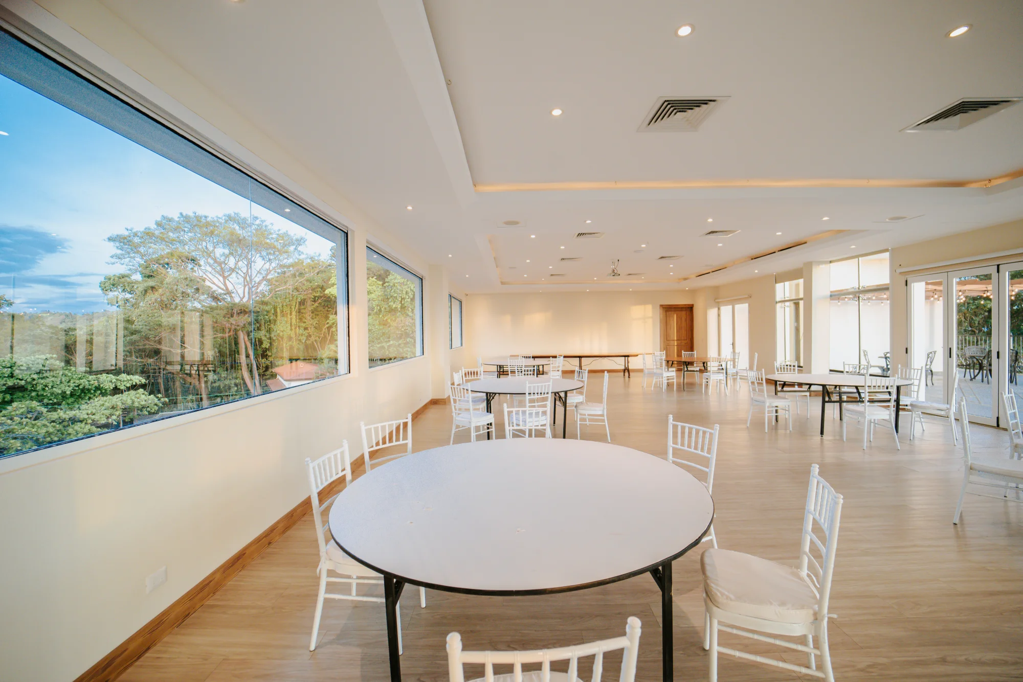Bright event room at Parador with a round table and white chairs by floor-to-ceiling windows looking out over the rainforest and the Pacific.