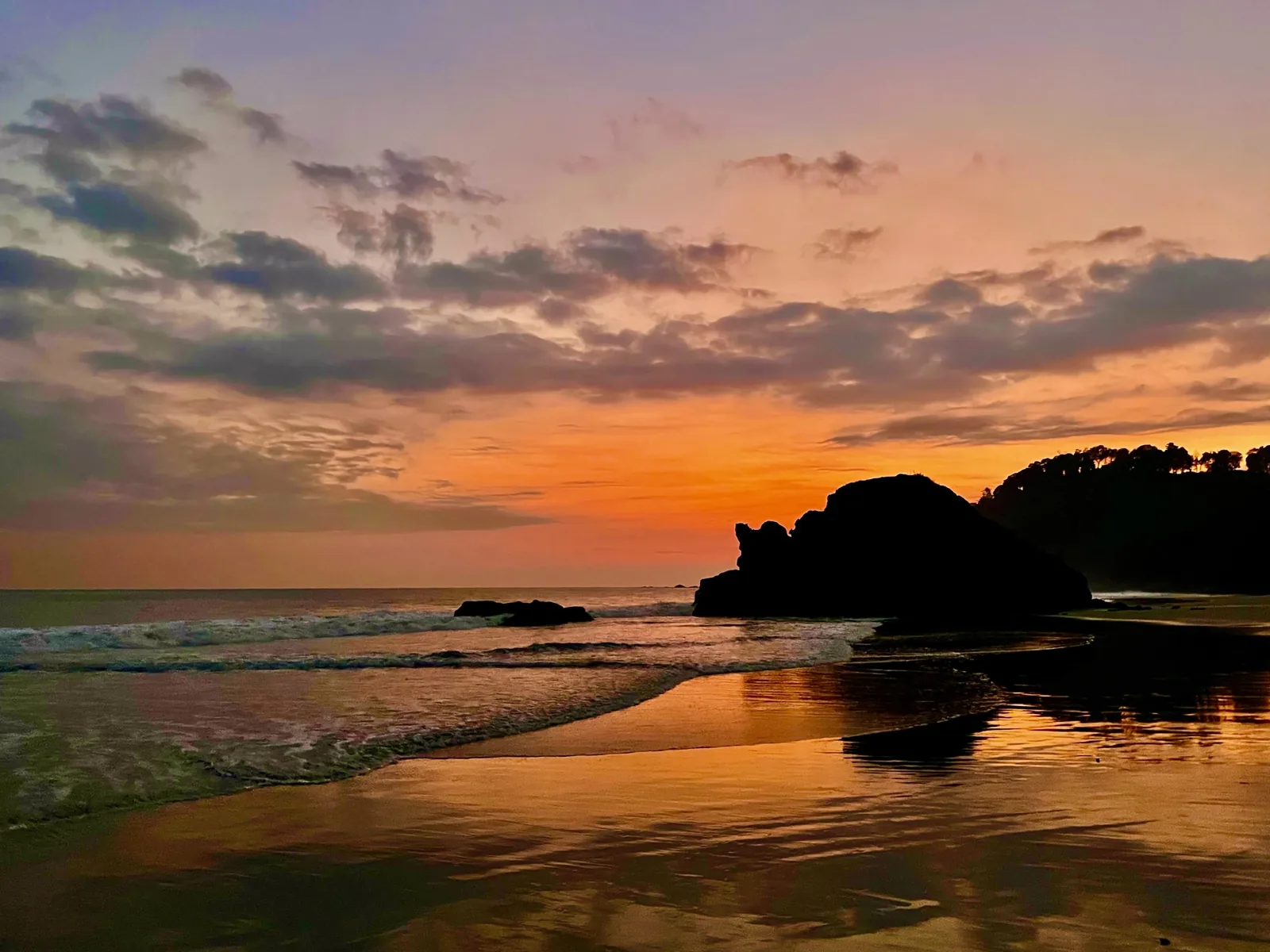 Un atardecer naranja y morado sobre el Pacífico, con una roca oscura recortada contra la arena mojada.