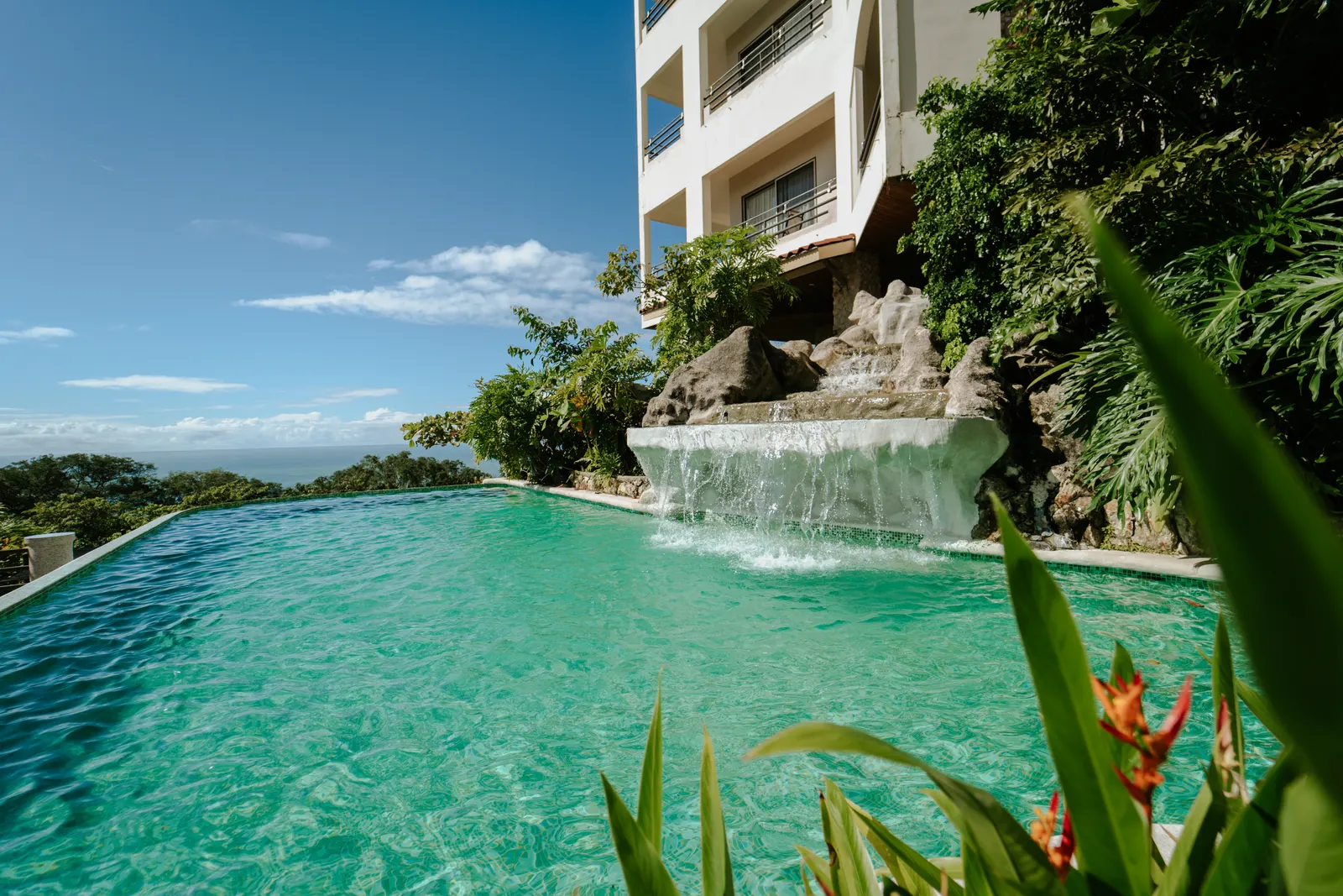 Una cascada de piedra alimenta una piscina infinita junto al edificio del hotel, con flores de heliconia en primer plano.