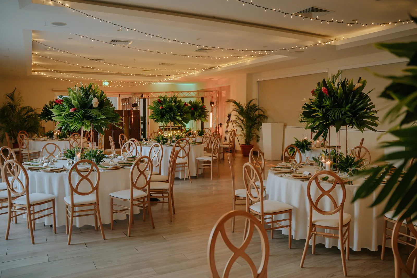 Salón de recepción con mesas redondas, altos centros de mesa de palma y jengibre rojo, sillas de madera con respaldo ovalado y luces colgantes en lo alto.