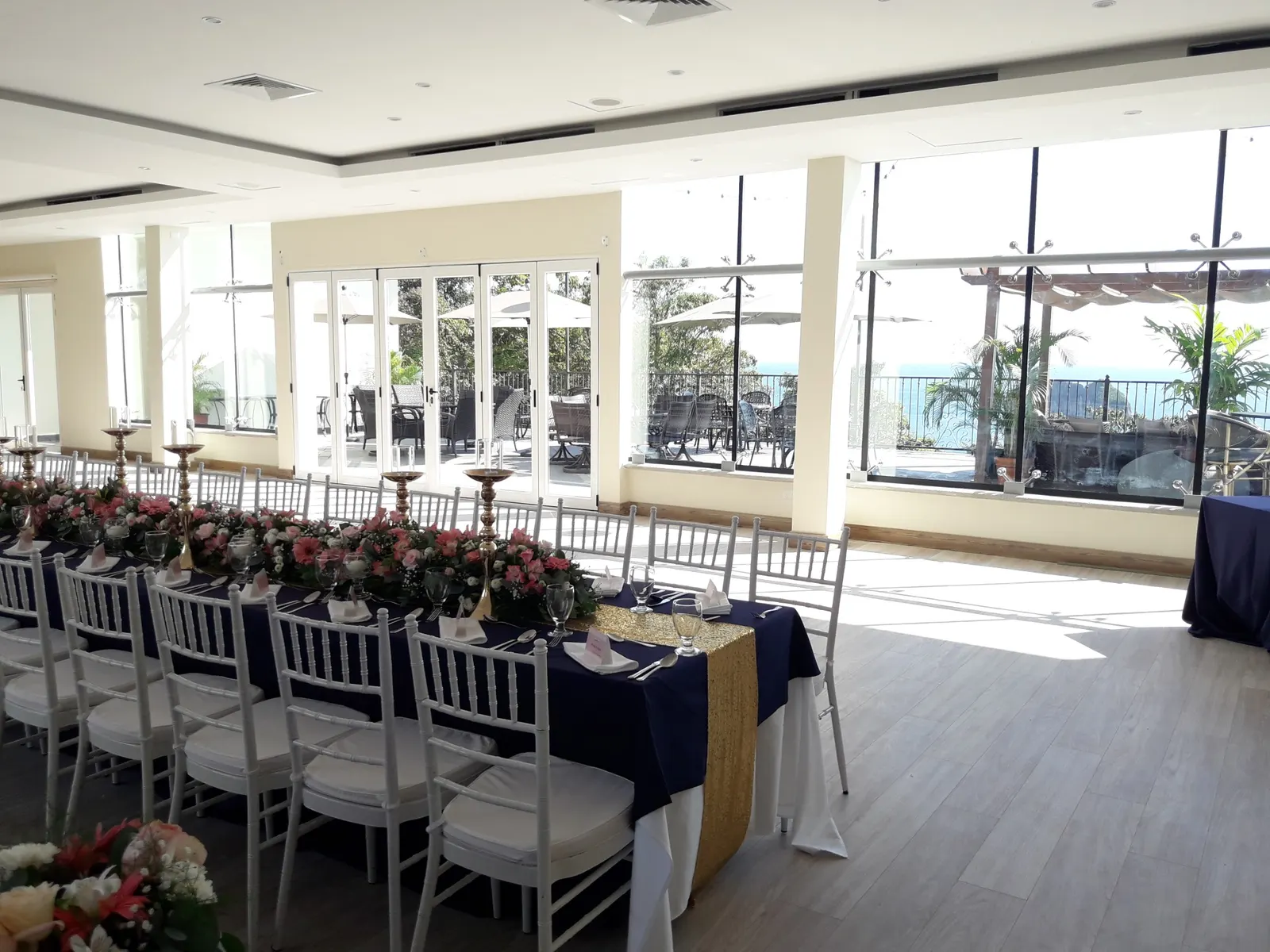 Long banquet table set with navy linens and a gold runner, glass doors open to the oceanview terrace.