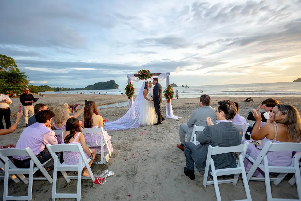 Invitados en sillas de una ceremonia en la playa aplauden mientras los recién casados están juntos bajo un arco floral al atardecer.