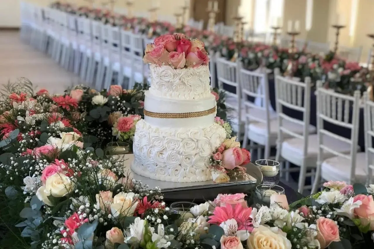 Pastel de bodas blanco de tres pisos con rosetas y una cinta dorada, coronado con rosas en tonos rosa pálido y rosado.