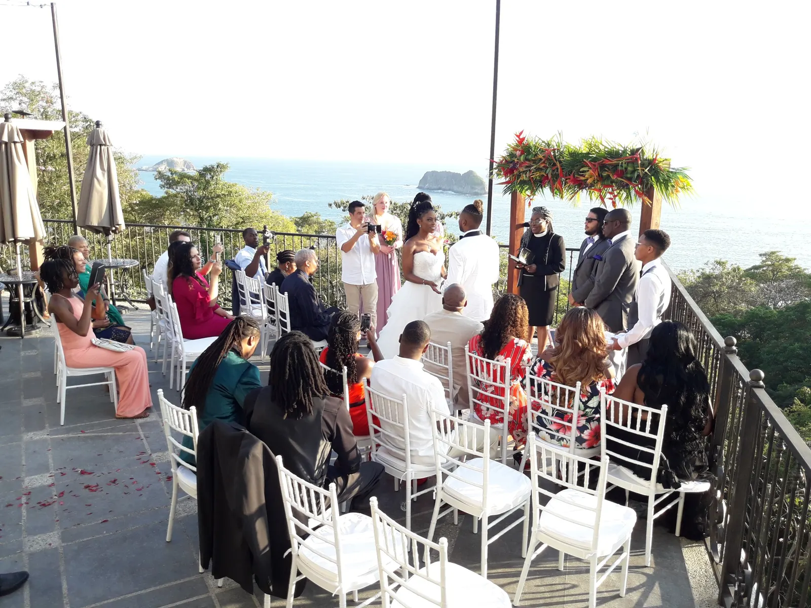 Ceremonia de boda al aire libre en la terraza bajo un arco cubierto de heliconias, con invitados sentados en sillas blancas e islotes visibles en el océano.