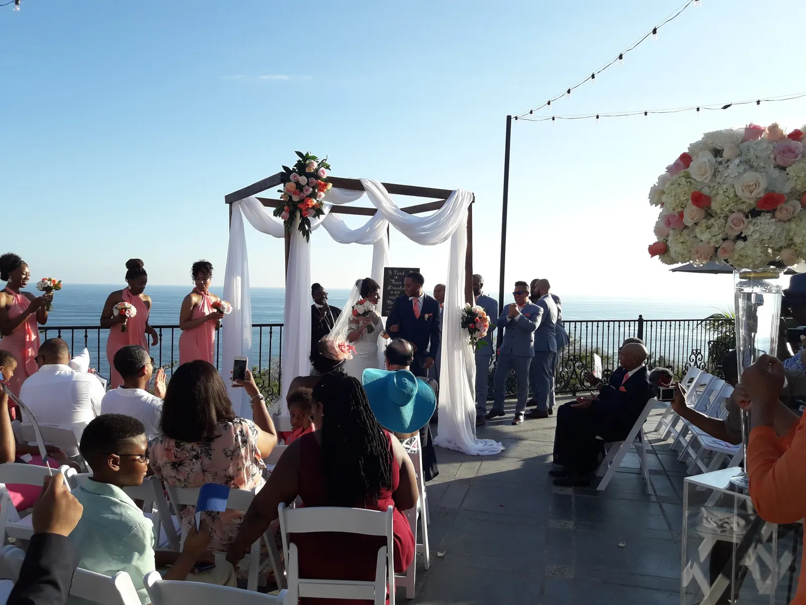 Ceremonia de boda al aire libre en una terraza con vista al océano, con damas de honor en vestidos color coral y padrinos en trajes grises junto a la pareja bajo un arco con telas blancas, y los invitados sentados aplaudiendo.