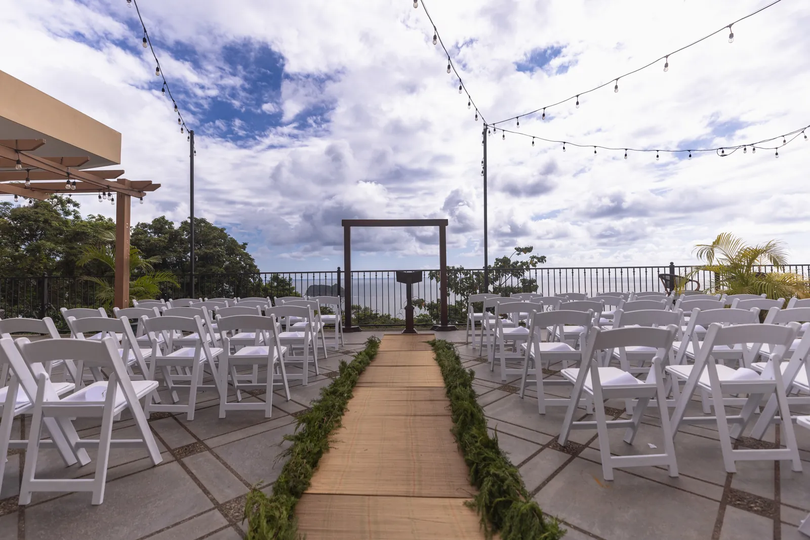 Espacio de ceremonia vacío en la terraza con filas de sillas blancas, una alfombra en el pasillo, un arco de madera y el océano detrás.