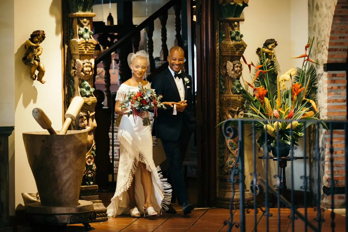 Los novios caminan juntos a través de una puerta de madera tallada decorada con flores de heliconia y estatuas de querubines.