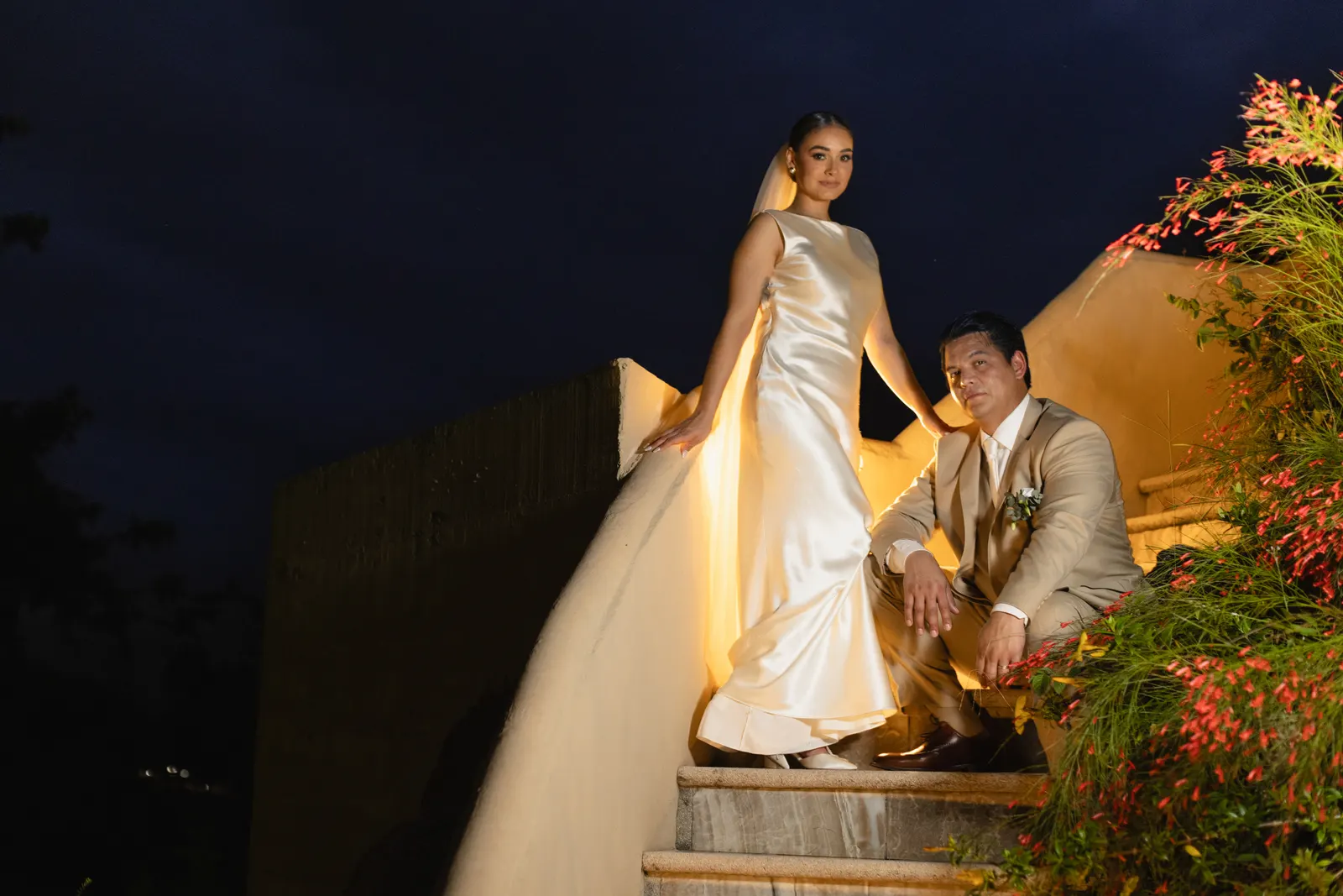 Una novia con un vestido de satén entallado de pie sobre unas gradas de piedra de noche, junto al novio sentado con traje color canela, con luz cálida y plantas en flor cerca.