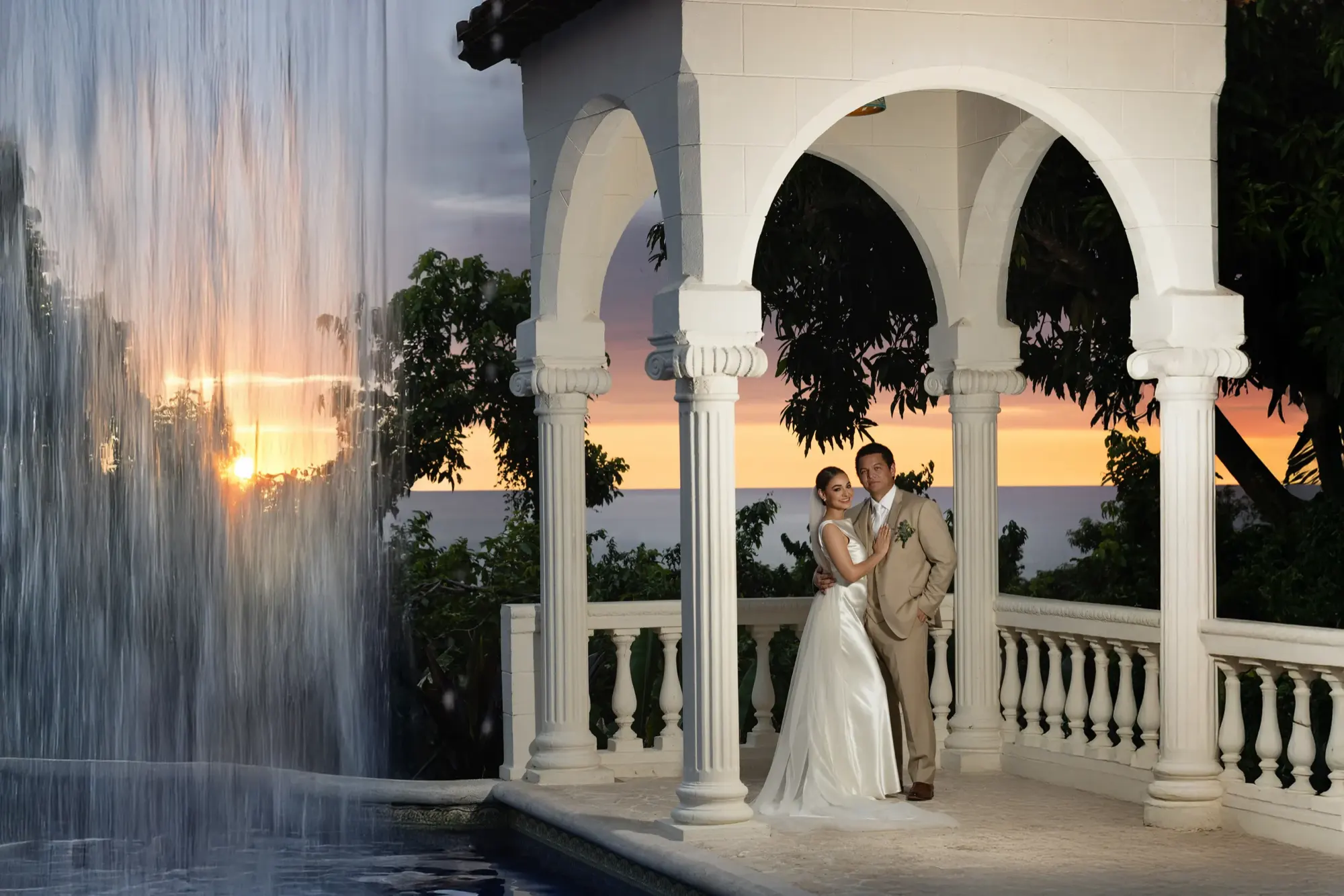 Vista amplia de una cascada y un gazebo en Parador, con la pareja de pie bajo los arcos mientras el sol se pone sobre el océano.