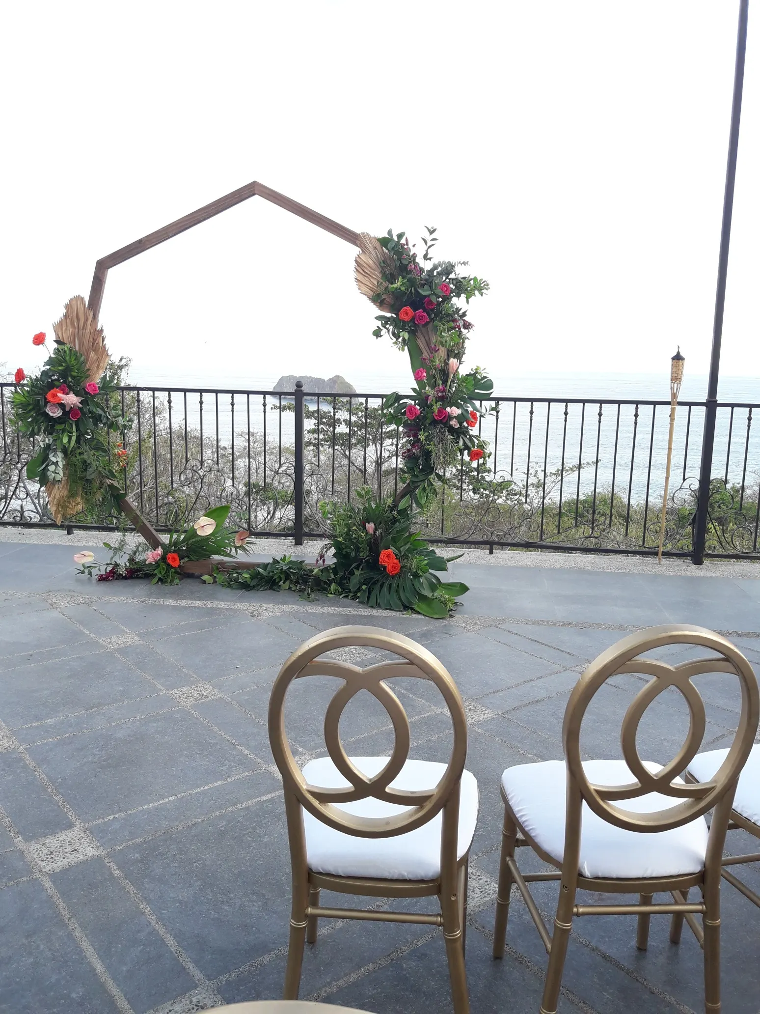 Arco geométrico de boda con rosas en cascada en una terraza con vista al mar, con sillas doradas frente al Pacífico.