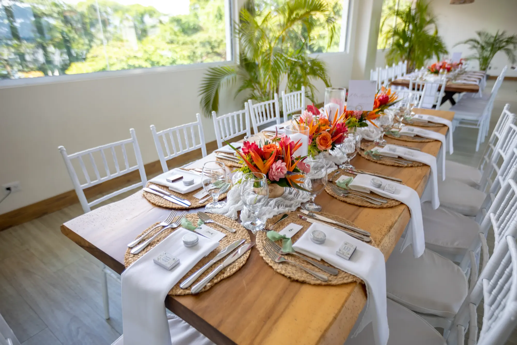Comedor de recepción con una mesa larga servida para los invitados, centros de mesa tropicales y ventanales de piso a techo.