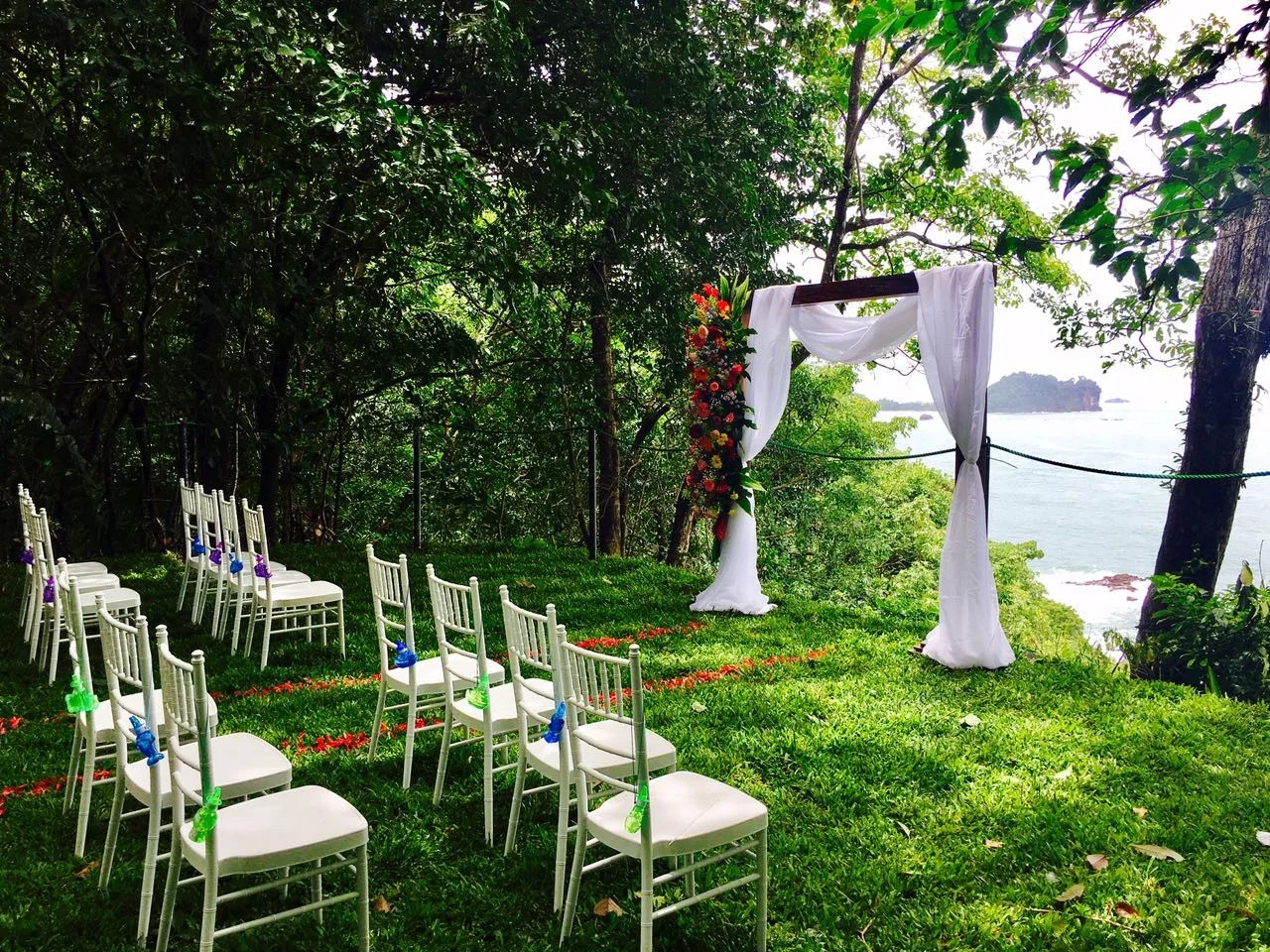 Césped de ceremonias vacío con vista al océano en Pacific Vista Point, con sillas chiavari blancas, un arco floral y un pasillo de pétalos enmarcado por árboles del bosque tropical.