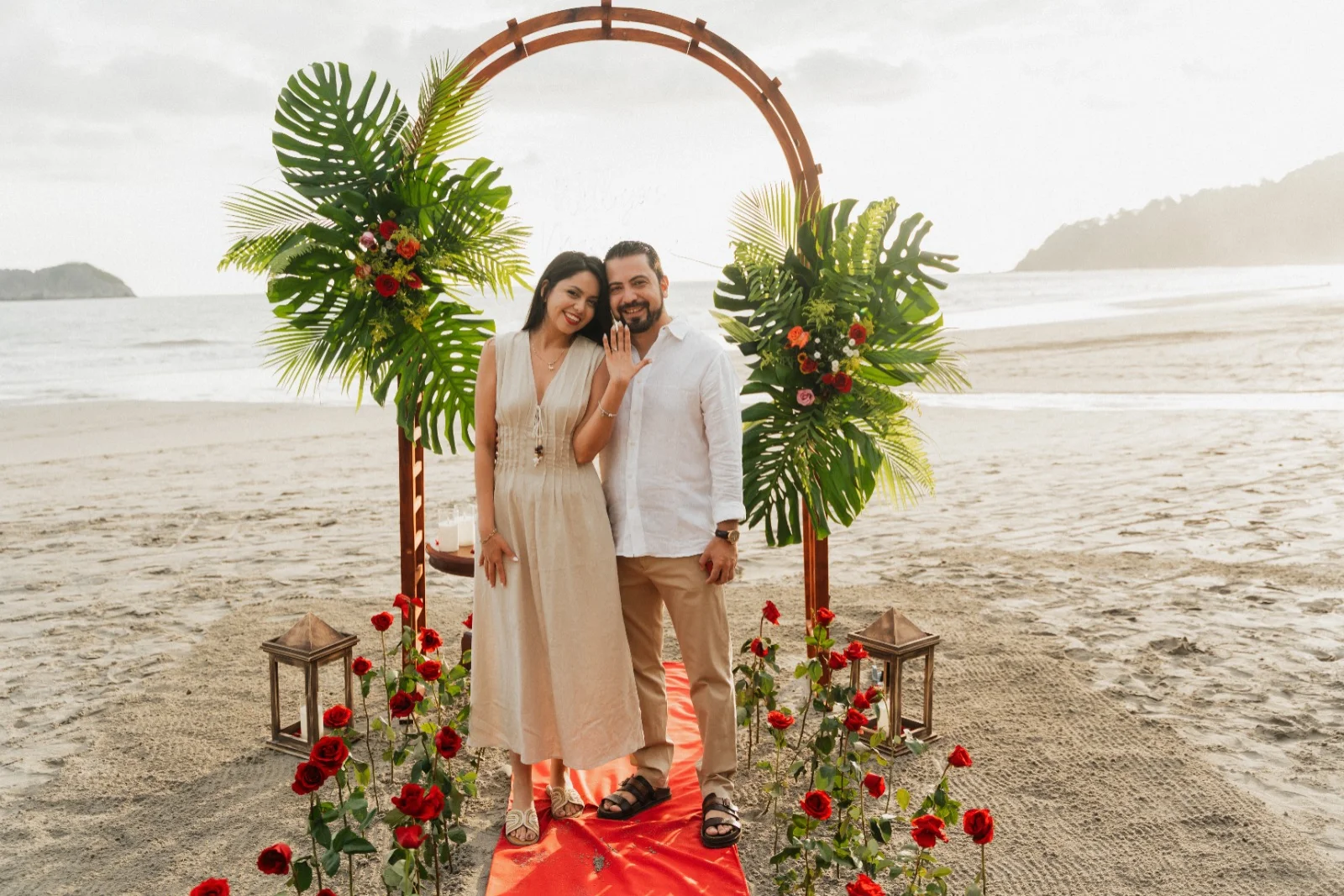 Una pareja se abraza bajo un arco floral decorado con hojas de palma y rosas rojas en la playa de Miamar al atardecer, con un pasillo de rosas a sus pies.